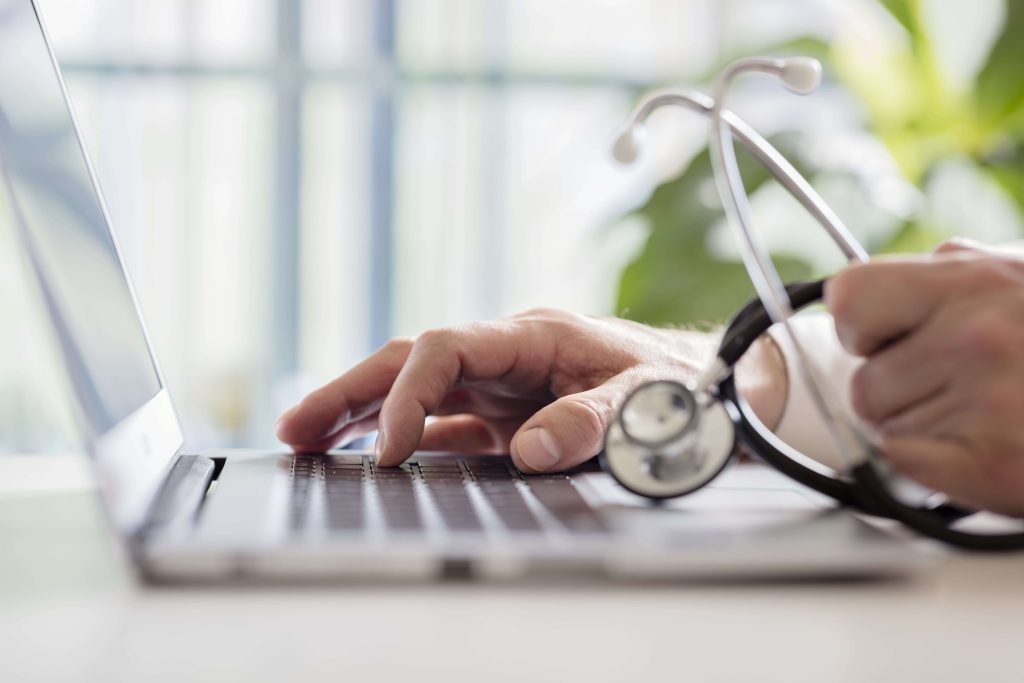 Hands typing on a laptop beside a stethoscope